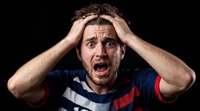 Male athlete clutching head and shouting, wearing navy jersey, intense emotional closeup