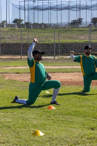 Male baseball teammates stretching during sunny warmup on green field with training cones