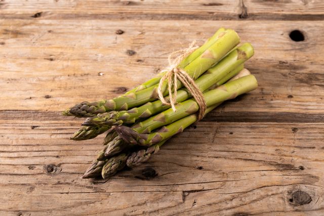 This image shows a bunch of fresh organic asparagus tied together with twine, placed on a rustic wooden table. Ideal for use in food blogs, healthy eating articles, farm-to-table promotions, and vegetarian or vegan recipe websites. The natural and unaltered look emphasizes the freshness and organic quality of the produce.