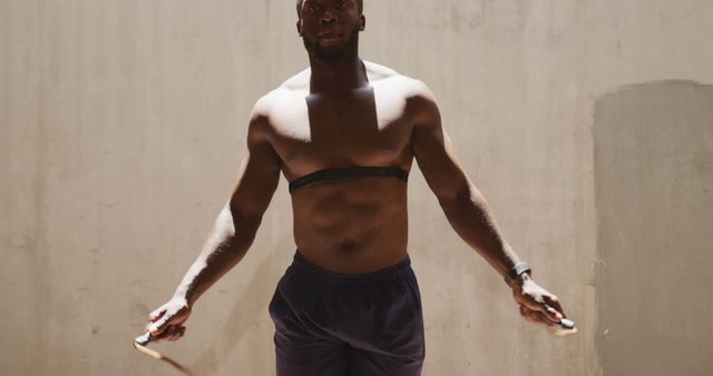 Front view of a shirtless African American man wearing a chest strap heart rate monitor and skipping with a rope in the shade under a bridge, while exercising on a sunny day in the city