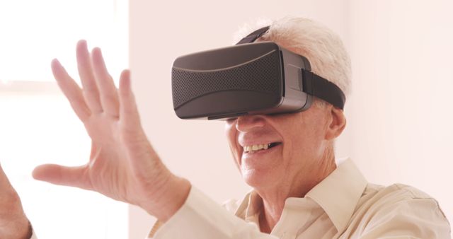 Senior man wearing a VR headset and smiling while interacting with virtual environment. Useful for depicting advancements in technology for older adults, showcasing joy and excitement in using virtual reality, or illustrating intergenerational tech experiences and innovation.