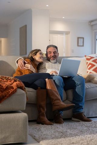 Mature couple cuddling on sofa while browsing laptop together in cozy living room with dog