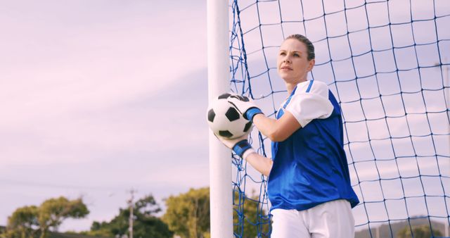 Female soccer player standing confidently next to goalpost while holding ball. Ideal for use in sports advertisements, team promotions, athletic wear, women’s empowerment in sports, and soccer training materials. Emphasizes strength, skill, and dedication in team sports.