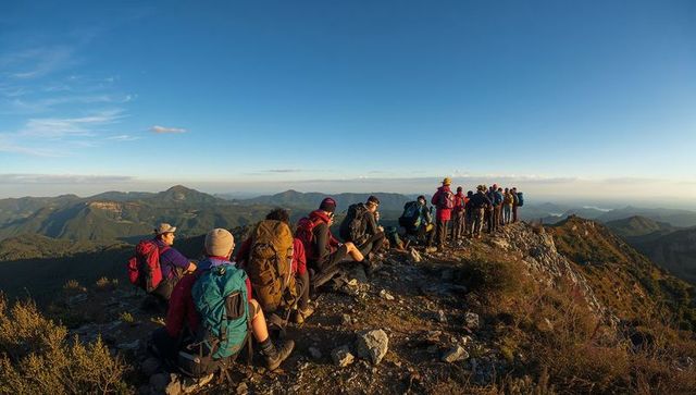 Hikers pausing along narrow rocky ridge at mountain summit during golden hour panorama