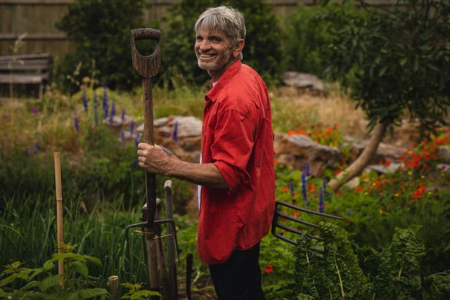 Senior man smiling while holding a pitchfork in a lush garden. Ideal for content related to gardening, sustainable living, rural lifestyle, retirement activities, and healthy hobbies. Can be used in articles, blogs, and advertisements promoting outdoor activities, gardening tips, and senior well-being.