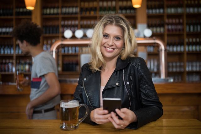 Portrait of smiling beautiful woman using mobile phone while having glass of beer in bar