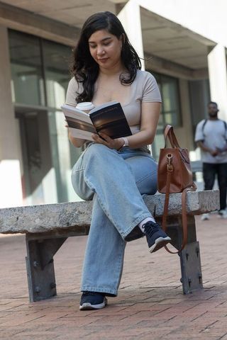 Young woman sitting on urban bench reading paperback with coffee and leather bag