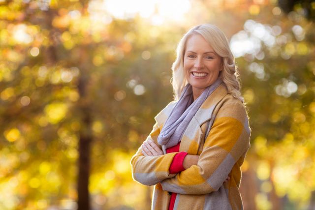 Woman standing in park during autumn with arms crossed, smiling warmly. Ideal for use in lifestyle blogs, seasonal promotions, outdoor activity advertisements, and wellness articles.