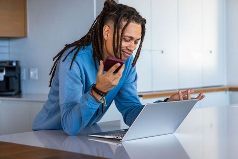 African american man leaning on kitchen island holding phone and checking laptop for remote work