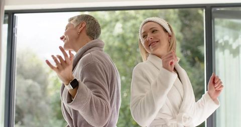 Two women dancing in plush bathrobes near glass doors, enjoying cozy morning at home