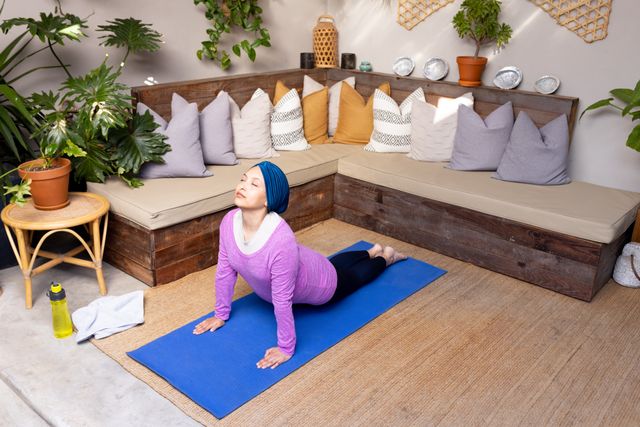 Relaxed biracial woman in hijab stretching in yoga pose on mat in living room, with copy space. Happiness, health, fitness, inclusivity and domestic life.