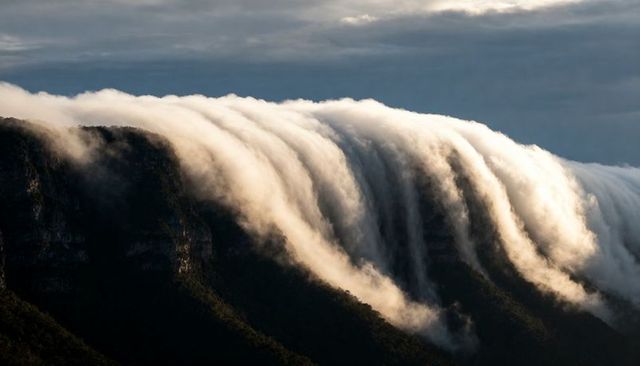 Sunlit cloud waterfall cascading over mountain ridge, dramatic misty sunrise scene