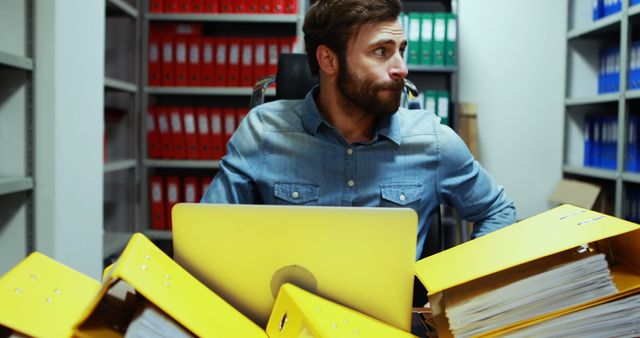 Depicts stressed office worker surrounded by yellow folders on desk, using laptop in modern office with files and shelves. Ideal for illustrating stress, workload, productivity issues, and office environments.