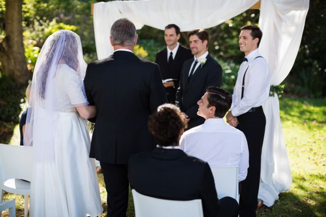 Bride and groom exchanging vows in front of a minister during an outdoor wedding ceremony. Guests are seated, witnessing the moment. Ideal for use in wedding planning websites, marriage blogs, event planning brochures, and romantic celebration promotions.