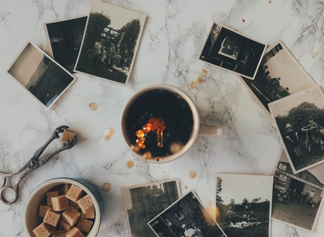 This visual depicts vintage black-and-white photographs scattered around a coffee cup on a marble table, emanating a sense of nostalgia and charm. Sugar cubes in a bowl and a sugar tong add to the vintage atmosphere. Ideal for blogs, articles, or advertisements focusing on nostalgia, history, vintage lifestyle, or coffee culture.