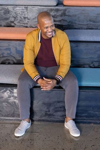 Smiling african american man sitting on tiered lounge bench wearing mustard bomber jacket