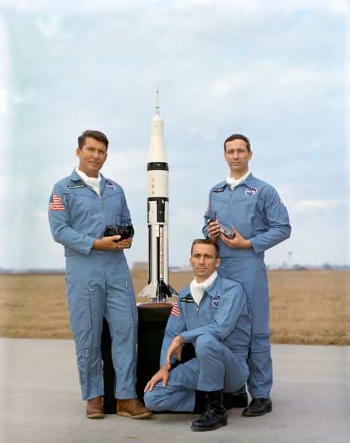 Three astronauts in blue NASA uniforms stand and kneel around a detailed model of a rocket on a clear day in 1968. The image features members of the Apollo 7 mission, a pivotal effort in the space race. The keywords 'Apollo 7,' '1968,' and 'NASA' highlight its historical relevance. This image can be used for educational materials, history books, articles on space exploration, and exhibitions related to space missions and NASA.