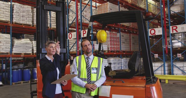 Warehouse manager holding clipboard discussing inventory with worker in safety vest near forklift. Boxes and pallets are visible on high shelves in background. Suitable for illustrating logistics, supply chain management, warehouse operations, and industrial employment scenarios.