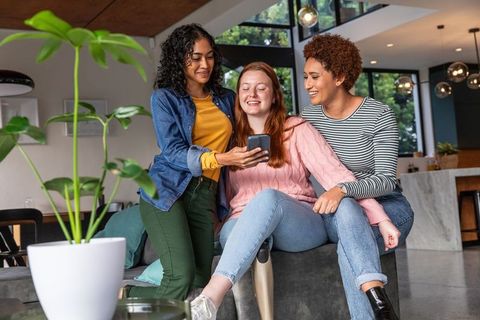 Diverse female friends sharing moment on smartphone in modern living room