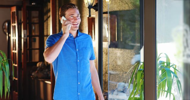 Man standing by a large window, talking on his phone and smiling, creating a warm and inviting scene. Ideal for themes related to communication, everyday life, home comfort, and indoor relaxation. Useful for advertisements involving telecommunications, lifestyle blogs, home appliances, and interior design.