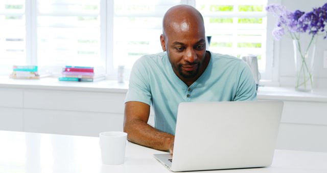 An African American man is intently using a laptop at a bright, modern home office. The setting includes a white desk, a cup, and a vase with purple flowers, adding a touch of color. This image can be great for content related to remote work, technology use, home office setups, or articles emphasizing productivity in a casual work environment.