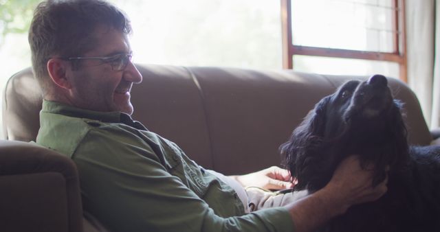 Middle-aged man sitting on sofa with black dog, enjoying a moment of bonding. Natural light from large window lighting cozy living room. Suitable for themes of domestic life, pet companionship, relaxation, leisure activities, and casual home lifestyles.