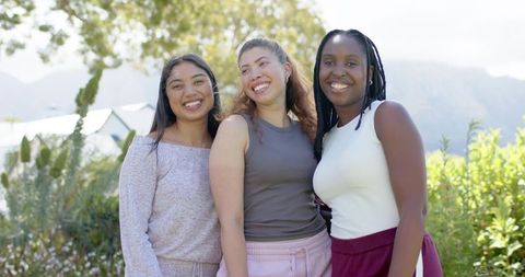 Three friends smiling and embracing in sunlit garden wearing casual summer outfits