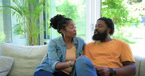 Black couple sharing calm conversation on sunlit sofa in living room, woman comforting man