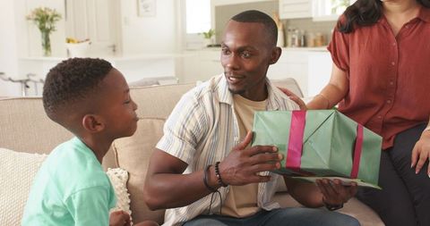 Father receiving wrapped gift from son on living room sofa celebrating warm family moment