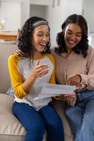 Diverse mother and daughter reading letter on sofa, sharing surprise and joyful family moment