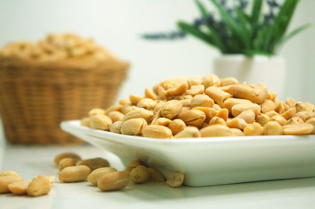 Roasted peanuts in a white ceramic plate suitable for use in health and food articles, blog posts about vegan diets, and nutrition websites. Can also be used for advertisements featuring healthy snacks, high-protein foods, and plant-based diets. The presence of a plant in the background adds a touch of rustic charm, making the image ideal for organic and natural product promotions.