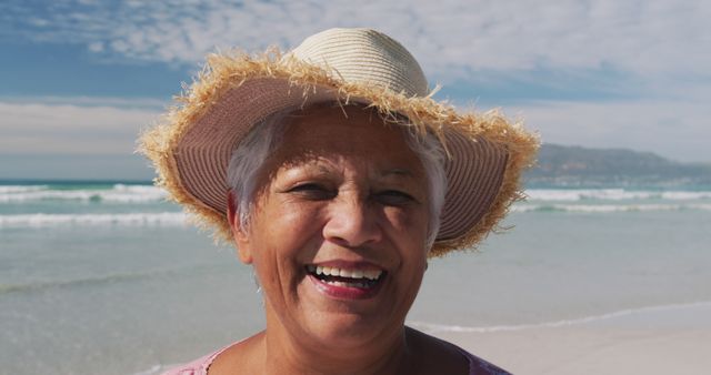 Senior woman enjoying sunny day at beach wearing a straw hat. Ideal for concepts related to joy, well-being, leisure, relaxation, and positive aging. Perfect for travel advertisements, wellness promotions, senior lifestyle magazines, and vacation websites highlighting happy and healthy retirements.