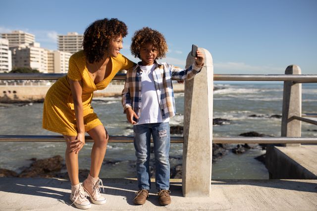 Mother and son enjoying a sunny day by the sea, standing on a promenade. The boy is using a smartphone to take a selfie of them. Ideal for use in family-oriented content, vacation promotions, and advertisements highlighting outdoor activities and family bonding.