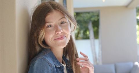 Sunlit relaxed young woman leaning on veranda wall wearing denim jacket showing ring
