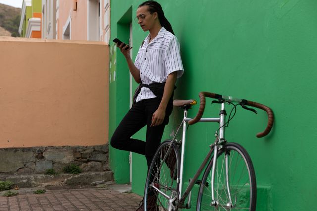 Trendy man with dreadlocks standing next to a green wall, using his smartphone with a bike beside him. Ideal for urban lifestyle, technology, and modern fashion concepts. Perfect for promoting mobile apps, cycling, or casual streetwear.