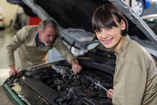Female mechanic smiling while examining a car engine in a repair shop. Ideal for use in advertisements, articles, and websites related to automotive services, gender diversity in the workplace, professional mechanics, and car maintenance tutorials.