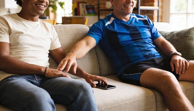 Male couple holding hands on sofa reaching for smartphone in sunlit living room showing casual intim
