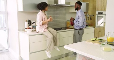 Couple chatting over morning coffee in sunlit modern kitchen, woman sitting on counter