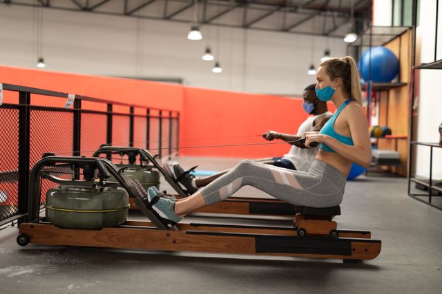 Caucasian woman and African American man working out on rowing machines in a gym while wearing face masks. This image can be used to promote fitness, health, and safety measures during the pandemic. It is ideal for articles, advertisements, and social media posts related to gym safety, workout routines, and maintaining an active lifestyle during health crises.