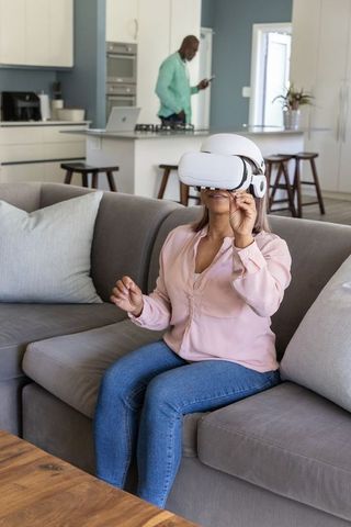 Diverse senior woman using VR headset on sofa while partner checks smartphone in kitchen