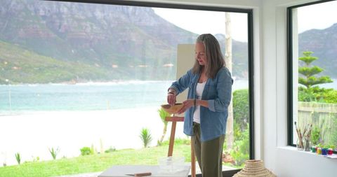 Senior woman mixing paints in bright coastal art studio overlooking sea