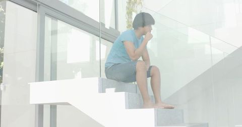 Pensive man sitting on floating white staircase by glass balustrade, foliage reflections