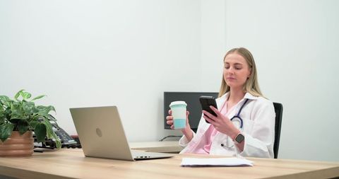 Female doctor checking smartphone while holding coffee at minimalist clinic desk