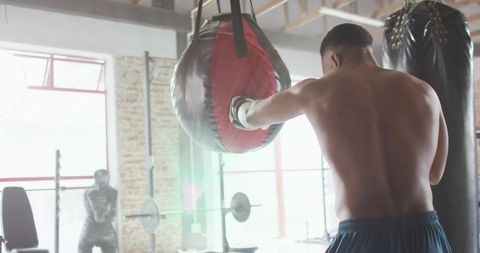 Shirtless boxer training with heavy bag in industrial gym with sunlight and lens flare