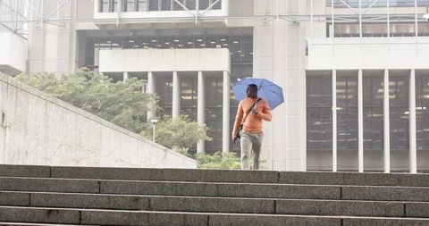African american man walking wet stairs with blue umbrella checking phone near office