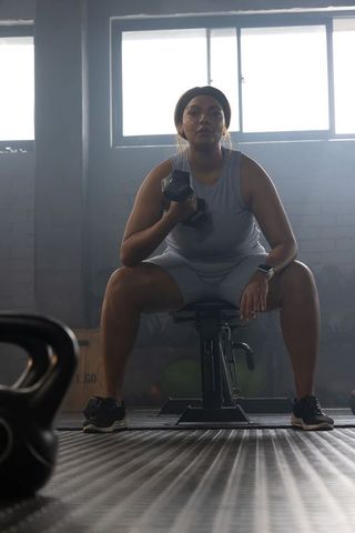 African american woman performing dumbbell seated curl on bench in moody backlit gym