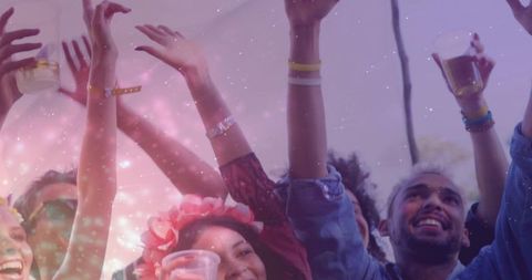 Festival crowd raising cups and cheering under canopy, friends wearing floral headband and wristband