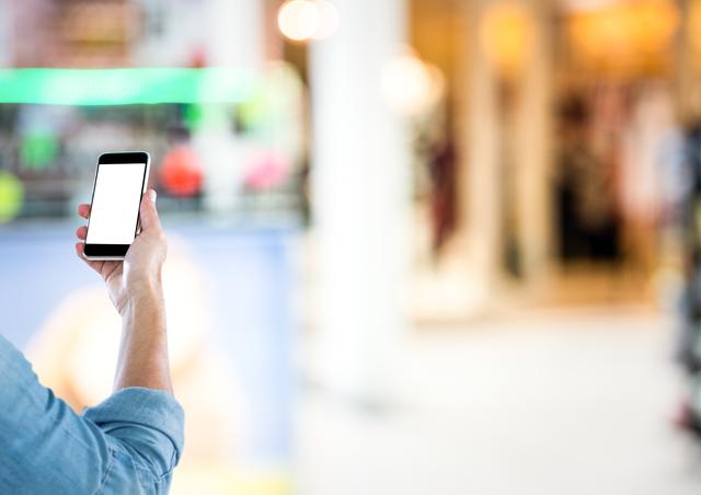 A person is holding a smartphone with a white screen, possibly browsing or using an app, in a retail store environment. The background is blurred, emphasizing the focus on the smartphone. Suitable for illustrating concepts related to mobile technology, shopping apps, consumer behavior, and modern retail experiences.