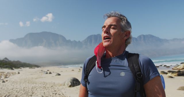 Senior hiker man with backpacks removing face mask and breathing while hiking on the beach. trekking, hiking, nature, activity, exploration, adventure concept.