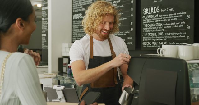 Barista with curly hair happily serving a customer at a trendy, modern cafe counter. The customer holds a phone while waiting for their order, creating a welcoming and engaging atmosphere. Ideal for use in promotional materials for cafes, ads illustrating customer service excellence, or articles about coffee culture and small businesses.